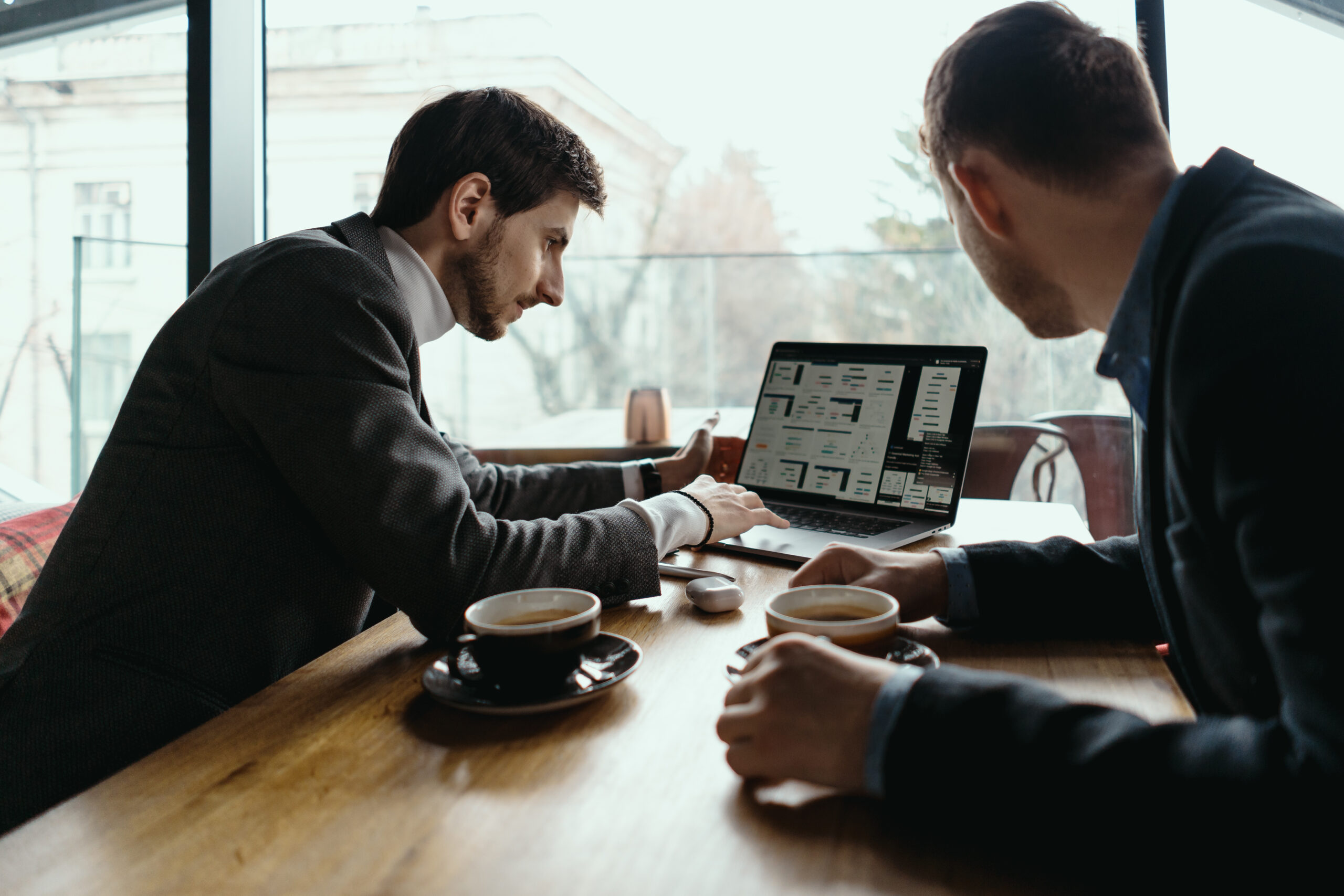 Two businessmen talking about new opportunities sitting with laptop at desk, planning project, considering business offer, sharing ideas while drinking coffee together